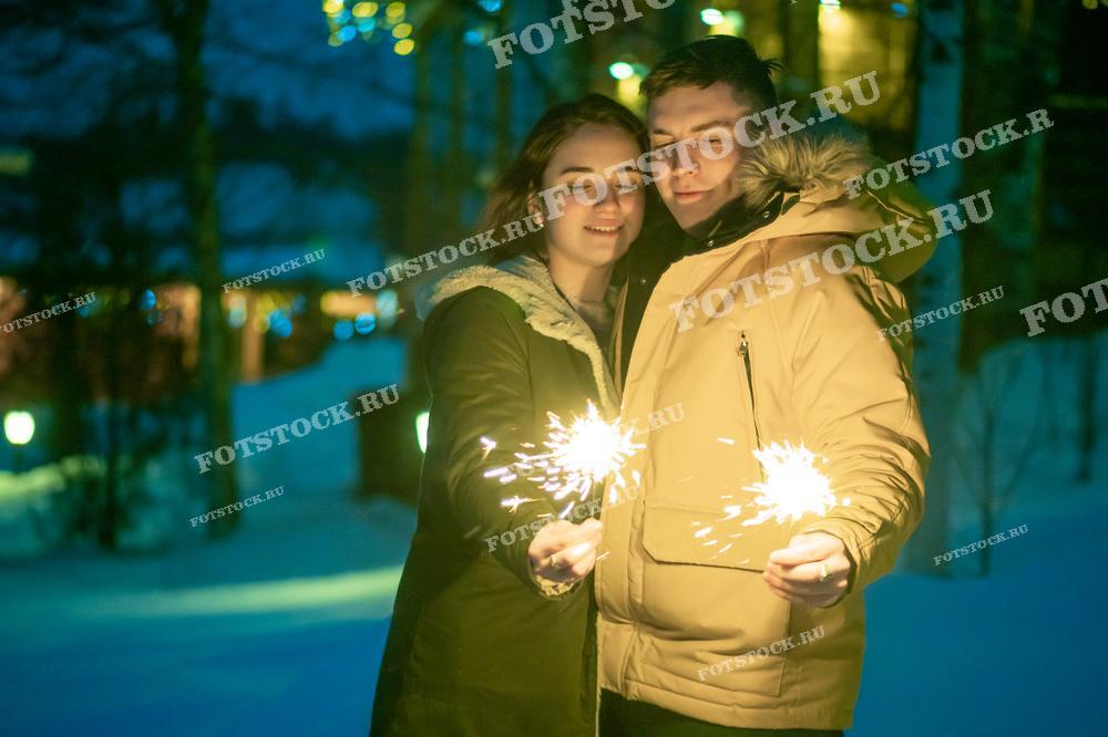 На фотографии изображена молодая пара, стоящая вместе на фоне вечернего зимнего пейзажа. Они одеты в теплую зимнюю одежду, а в руках держат зажженные бенгальские огни. Атмосфера наполнена праздником и романтикой.